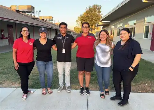 Six school staff members stand together outdoors on a school campus, smiling for a group photo.