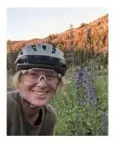 Woman in bike helmet leans into left side of photo with sunset bathed slope and purple flowers in background