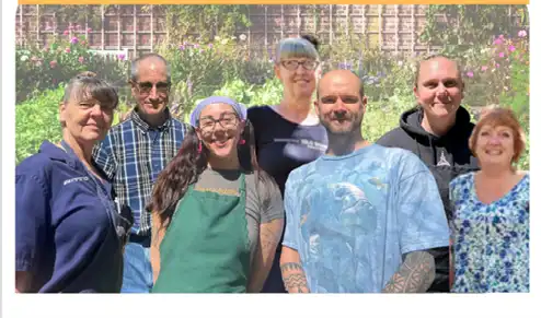 Seven individuals smiling in the sunshine with a garden faded in the background