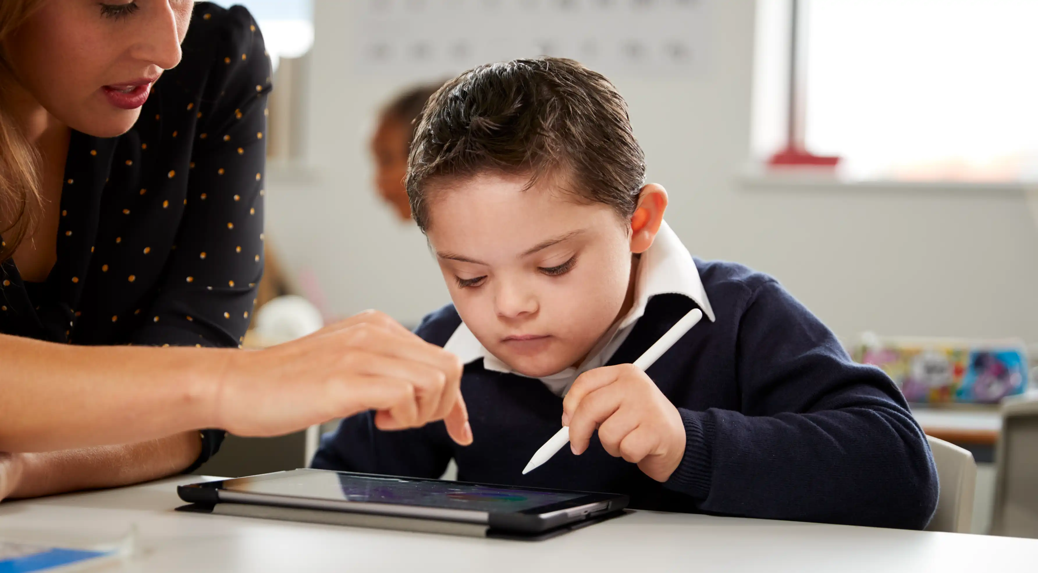 A child using an electronic pencil on an electric tablet while a teacher helps.