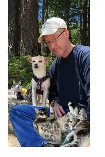 Adult in a ball cap sitting on a boulder in a forest with a small harnessed dog. There are three cats photoshopped on his lap and the boulder.