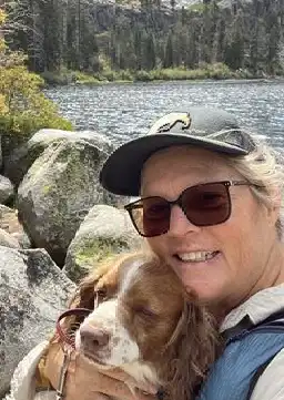 Headshot of a person in a cap and glasses holding a dog in front of an alpine lake