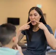 Woman pointing both hands at her lips while younger child is facing her and watching her.