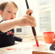 Young child holding paint brush with paint on it and trying to paint a piece of paper that is on a table.