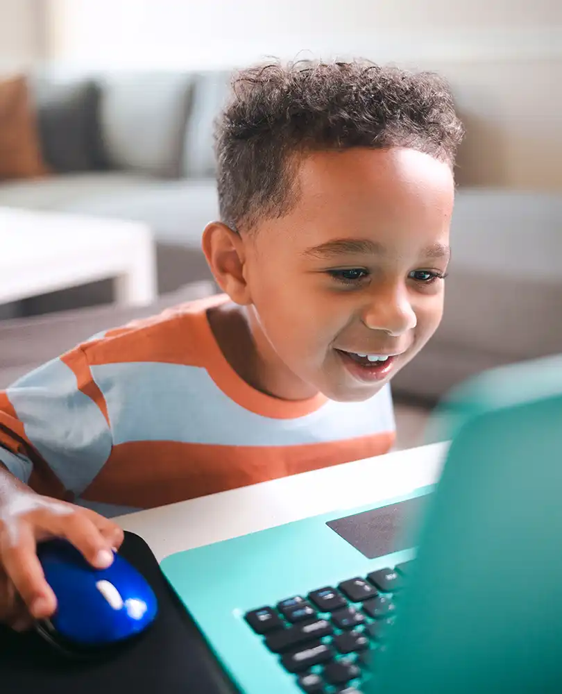 A photo of a young boy smiling while he operates a laptop computer.