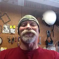 Faceshot from below of a white-bearded and mustached adult in front of a workshop wall
