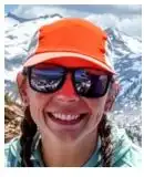 Smiling woman with braided pigtails and mirrored sunglasses and orange-billed cap with a snowy-alpine backdrop