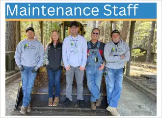 Five adults lean against the bucket of a tractor wearing jeans and Sly Park gear.
