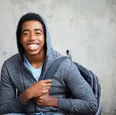 Teen wearing hooded sweatshirt while smiling at the camera and holding straps of a backpack
