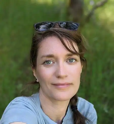 Portrait shot of a brown-haired woman with sunglasses perched on her brown hair smiling in front of blurred out grassy background