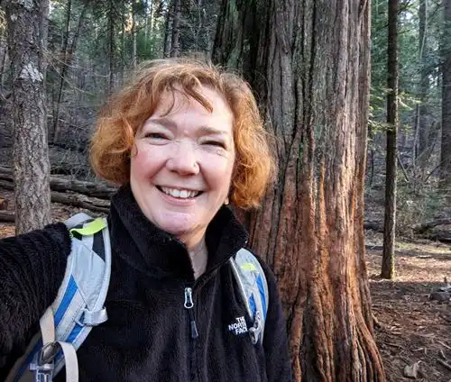 Smiling person with backpack on in front of cedar cedar tree