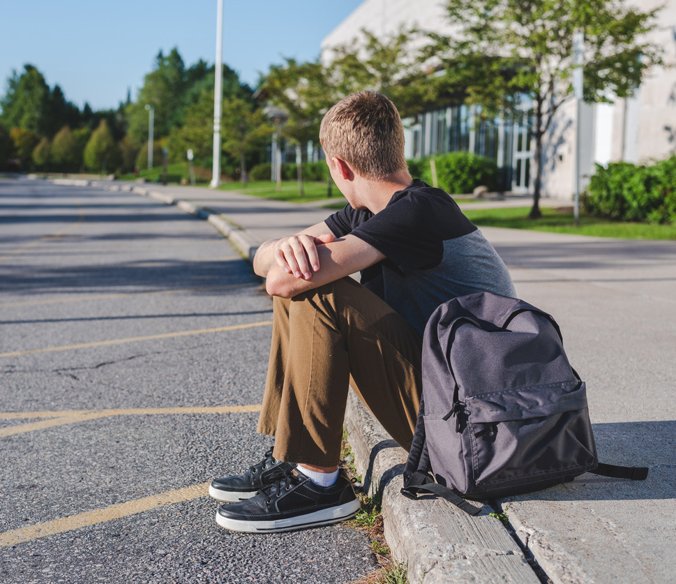 Photo of teen student sitting on a curb outside of school waiting for a ride.
