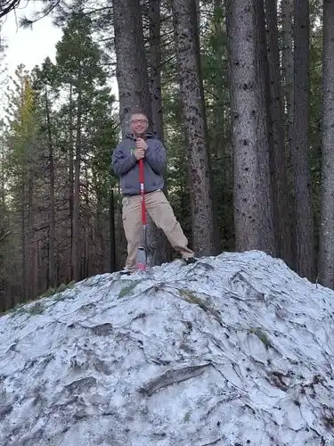 Person with a red-handled shovel standing atop a pile of dirty snow with conifers in the background.