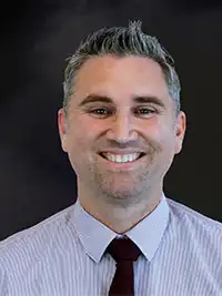 A smiling man with gray hair wearing a striped shirt and red tie in front of a dark gray background.